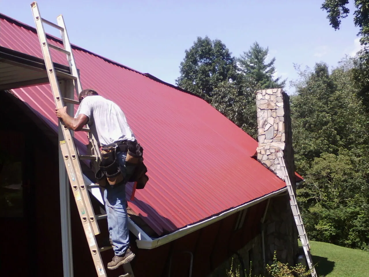 Roofer on a ladder beside a red metal roof and stone chimney — Soffit & Fascia Repair by Roofing Brothers in Scotrun
